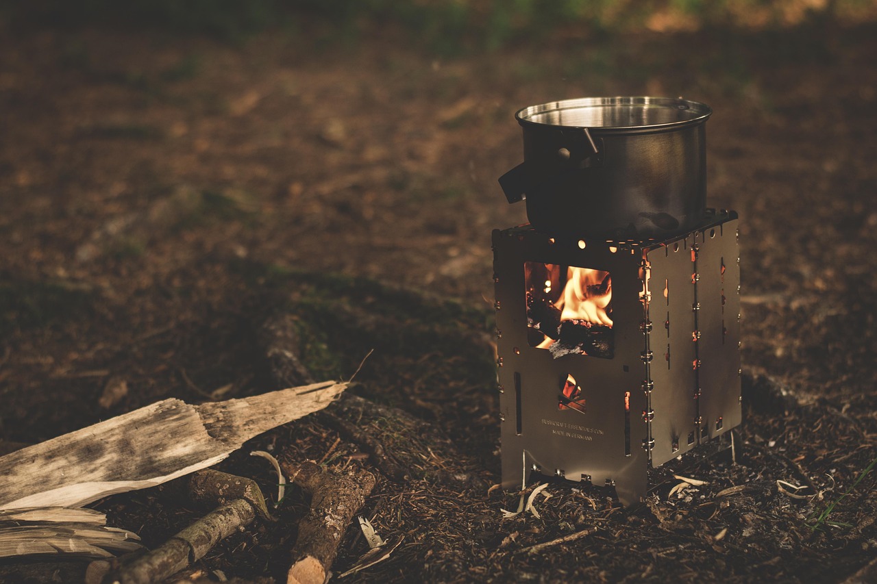 A metal camping pot heating on a small wood-burning stove outdoors at dusk