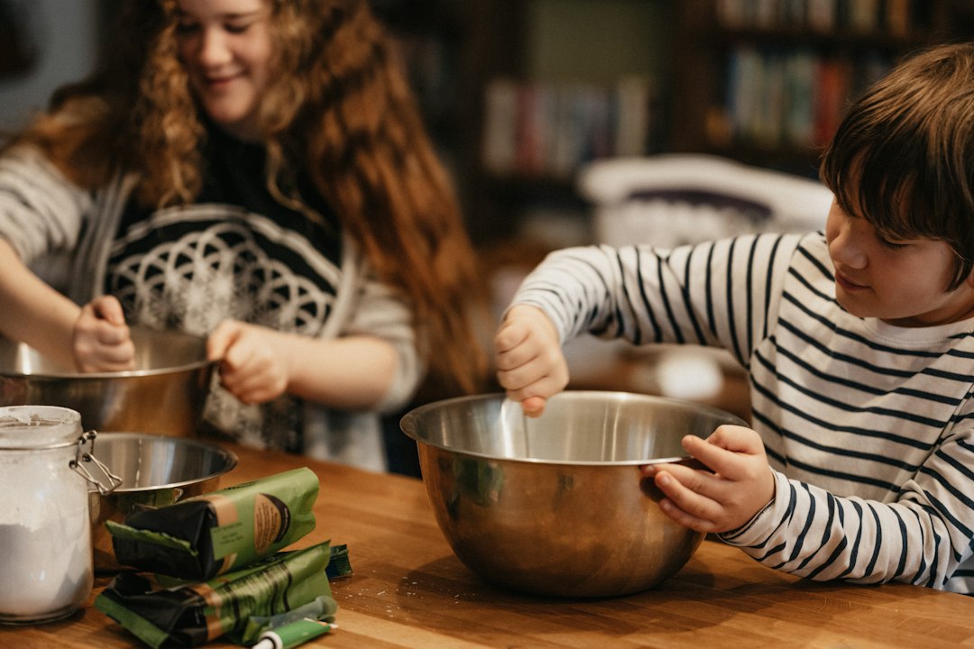 Two children at a wooden table stirring ingredients in metal mixing bowls